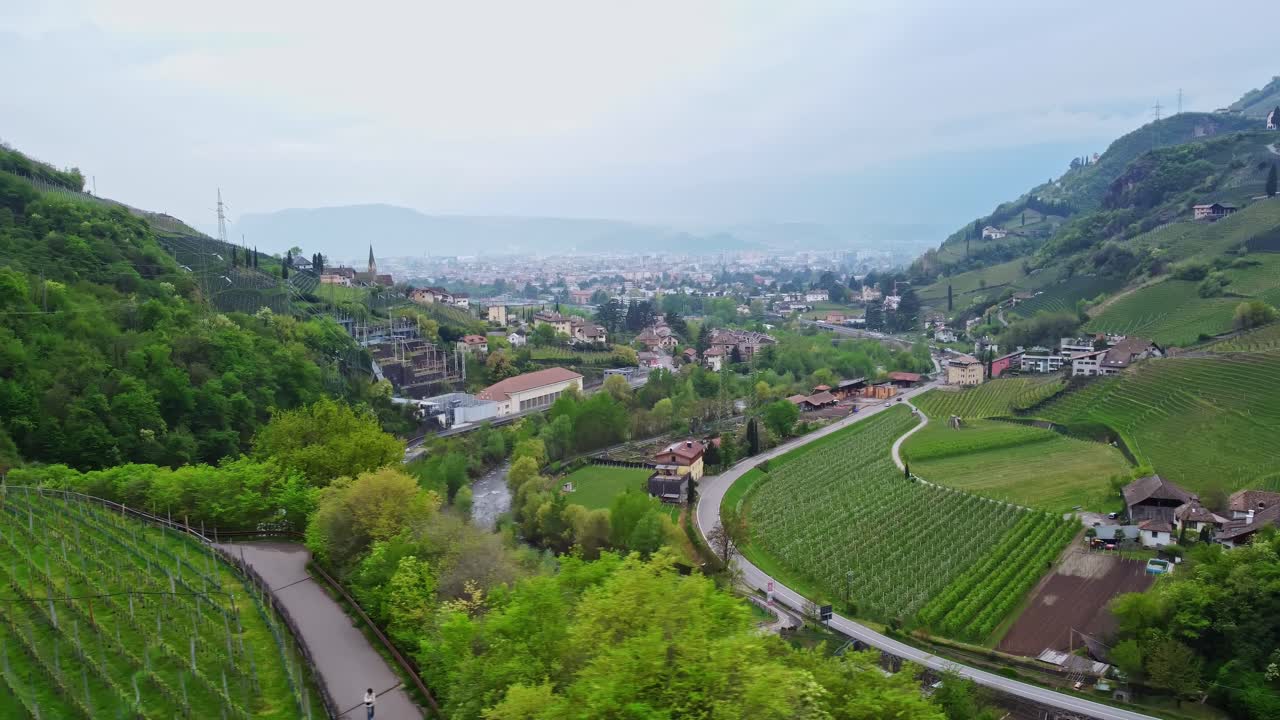 Drone view of green vineyards, a winding river, and street traffic near Roncolo Castle