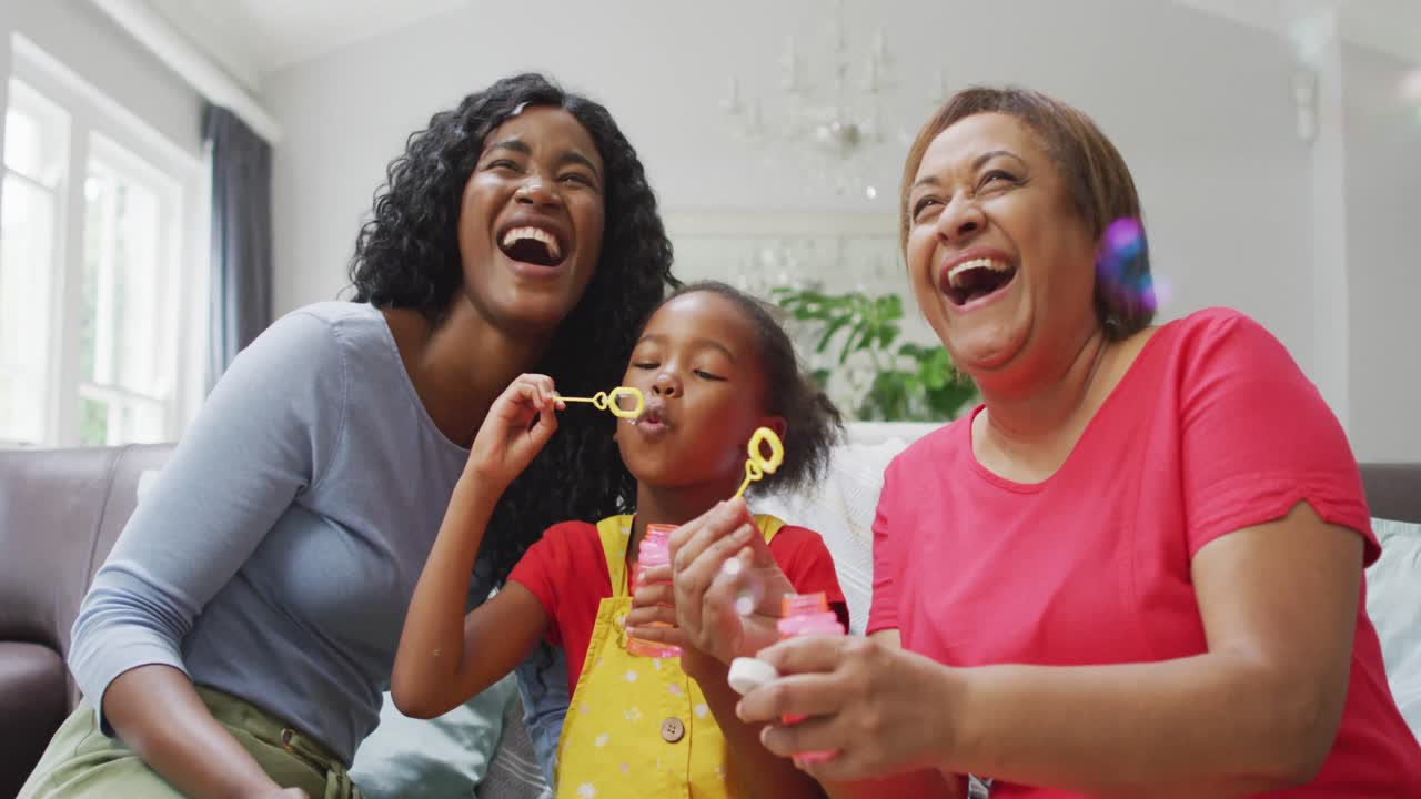 Happy african american grandmother, mother and daughter blowing bubbles at home