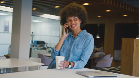 Smiling-Businesswoman-Standing-In-Modern-Open-Plan-Office-Talking-On-Mobile-Phone