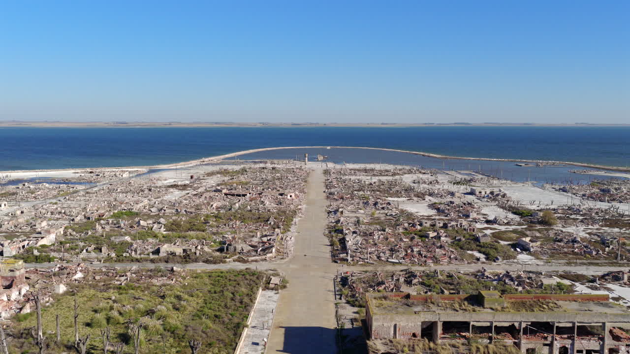 Fluido movimento aereo in avanti che segue un viale in rovina verso il blu profondo della Laguna Epecuén, evidenziando le croste minerali bianche e il paesaggio apocalittico, Pampas Argentinas, Buenos Aires