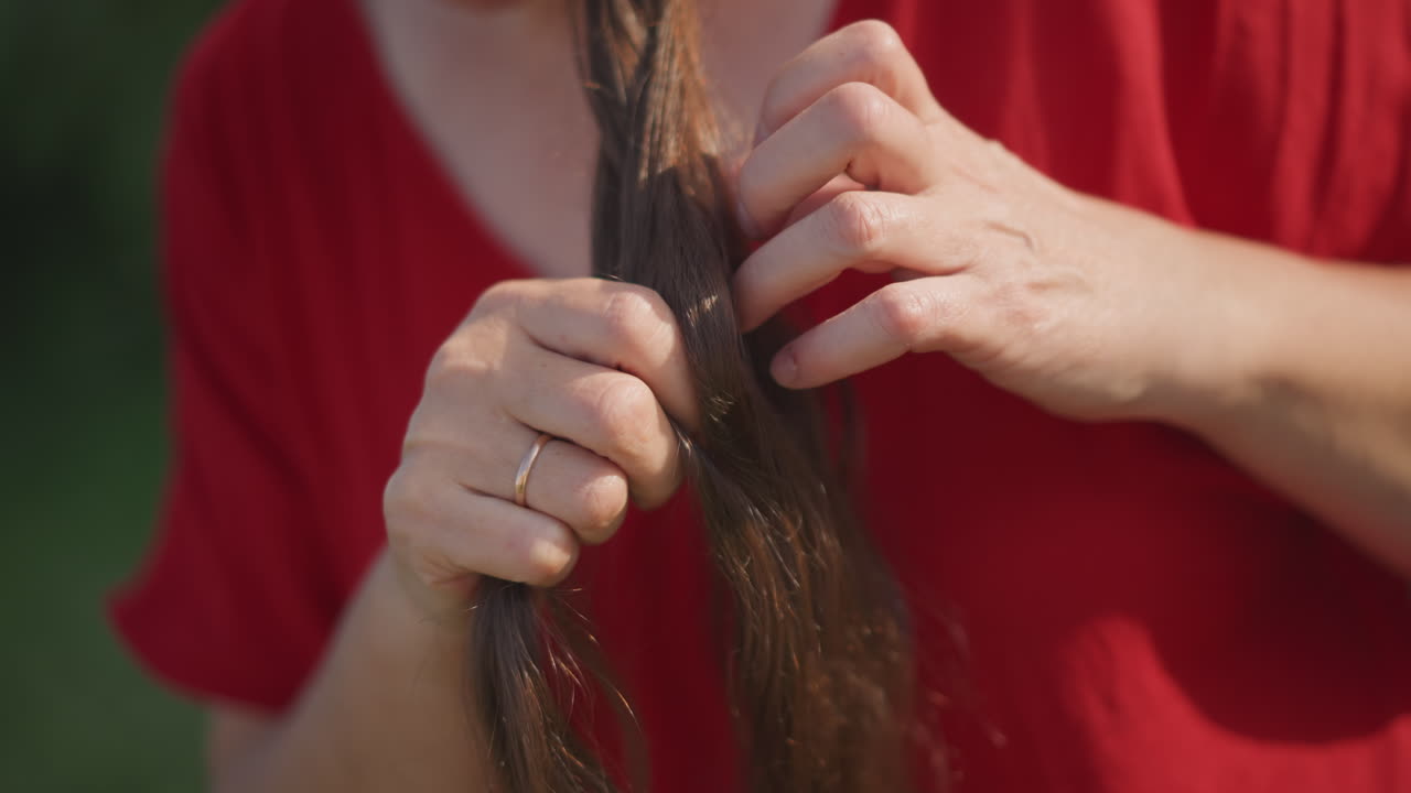 Wanita kaukasia mengepang rambut dari dekat, tangan fokus menjalin untaian dengan jari-jari mantap, penata rambut rumahan mempraktikkan teknik tepat di halaman belakang yang disinari matahari, suasana konsentrasi dan rutinitas hati-hati