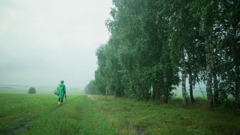 back view of woman in green raincoat carrying mint-colored bag and umbrella, wearing yellow rain boots, walking along muddy path in the distance surrounded by tall trees, greenery, and misty sky