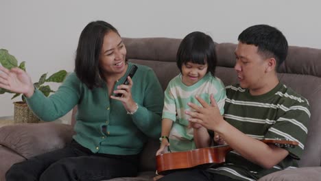 young happy asian family singing and dancing together while sitting on couch at home