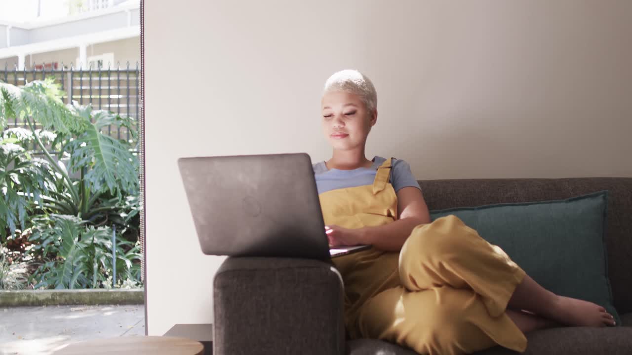 Happy biracial woman using laptop and sitting on couch in slow motion