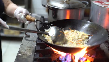 Chef-wearing-black-uniform-and-white-sterile-gloves,-skillfully-cooking,-mixing,-stirring-and-tossing-chao-fan-or-Chinese-fried-rice-in-a-wok-using-a-deep-ladle-on-full-blast-fire-in-a-kitchen