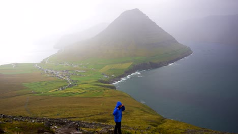 man wearing a blue jacket covers himself up and looks at malinsfjall faroese landscape, cold weather, wide