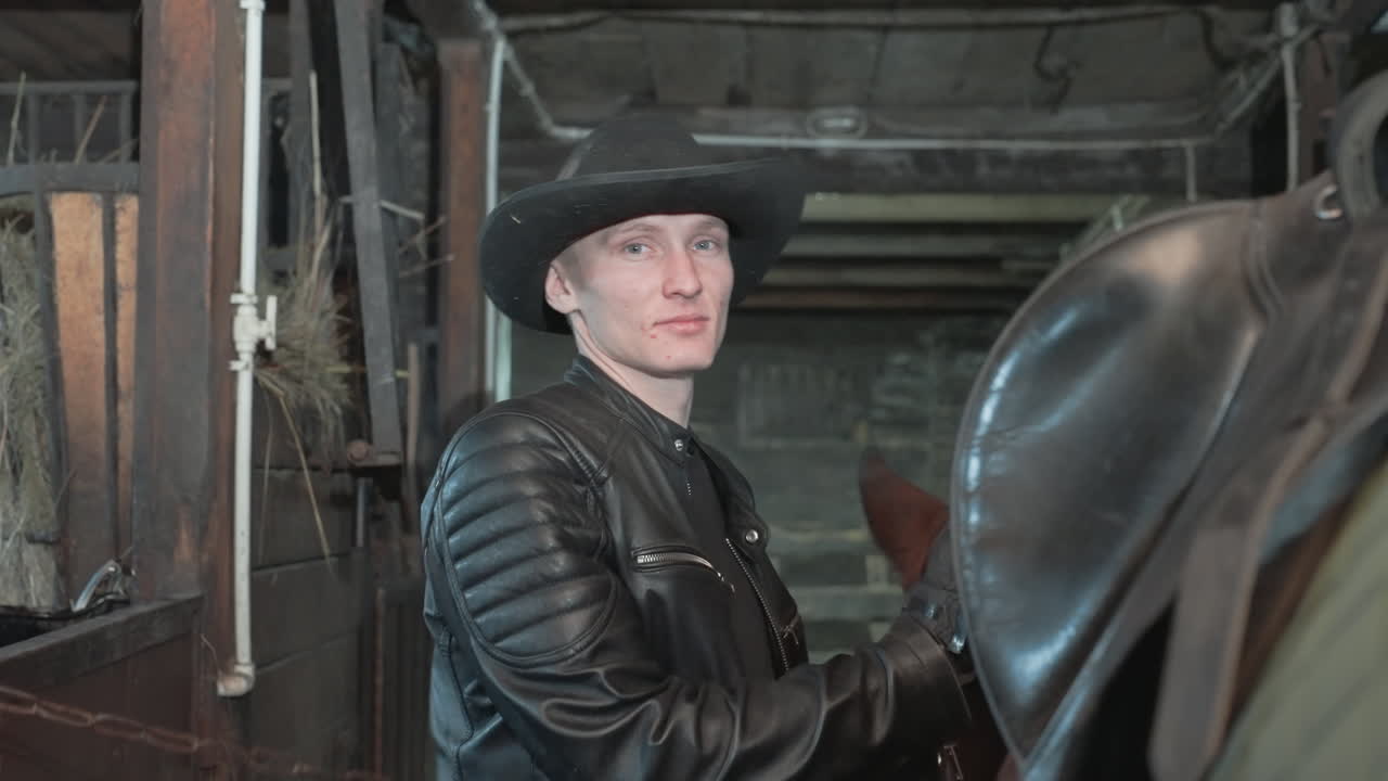 Uomo caucasico che sella un cavallo in una stalla, regolazione ravvicinata della sella e ispezione delle fibbie, finimenti in cuoio, espressione concentrata, giacca trapuntata e cappello, preparazione professionale per la cavalcata nel tranquillo corridoio del fienile.