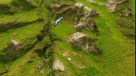 aerial birdseye view of a paraglider with a green mustached canopy sailing over bright green grass and rocky ravines