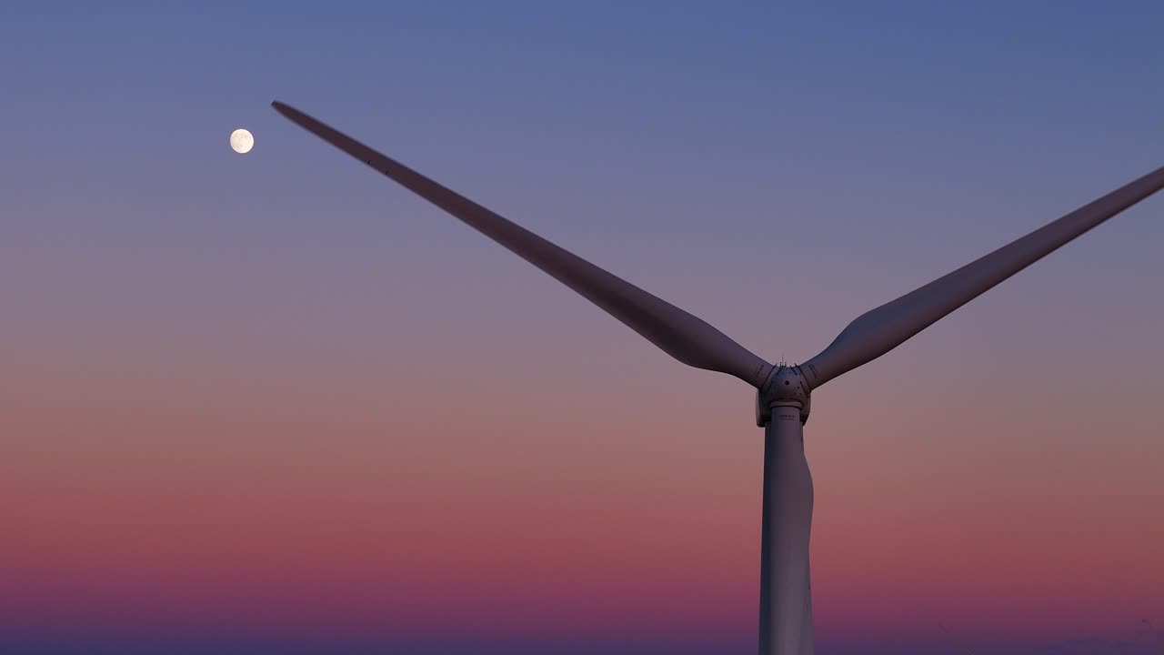 Vindturbinblader spinner og skaper fornybar energi under en måne på en gradienthimmel.