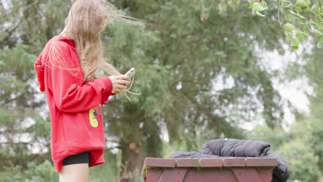 Next-to-wooden-bench-outdoors-young-girl-stands-using-her-smartphone