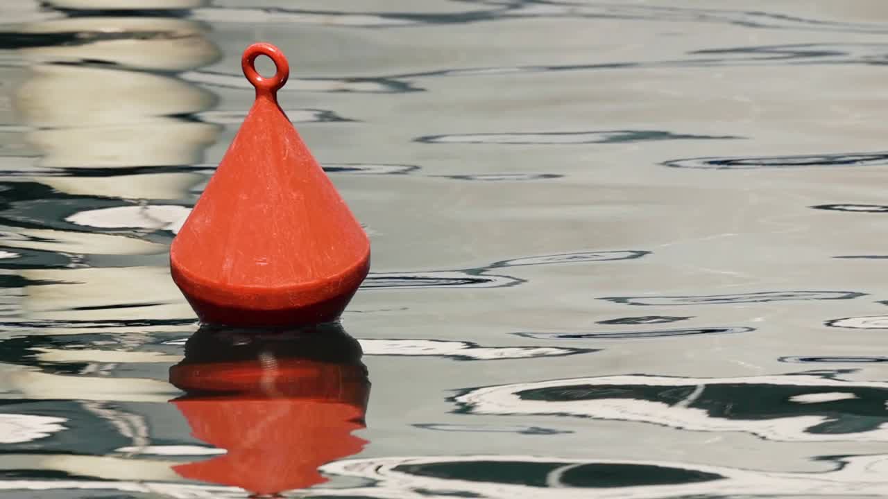 A bright red conical buoy bobs gently on calm, rippling water with a clear reflection