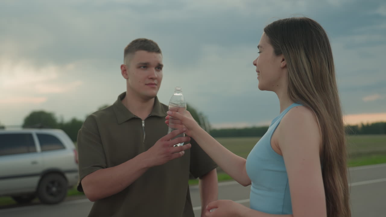 young couple paused on rural road as wife drinks water from plastic bottle then offers bottle to husband while husband watches attentively under cloudy sky beside parked car on quiet asphalt stretch