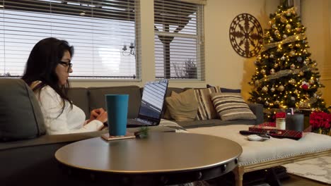 hispanic female working on laptop, on couch in front of christmas tree