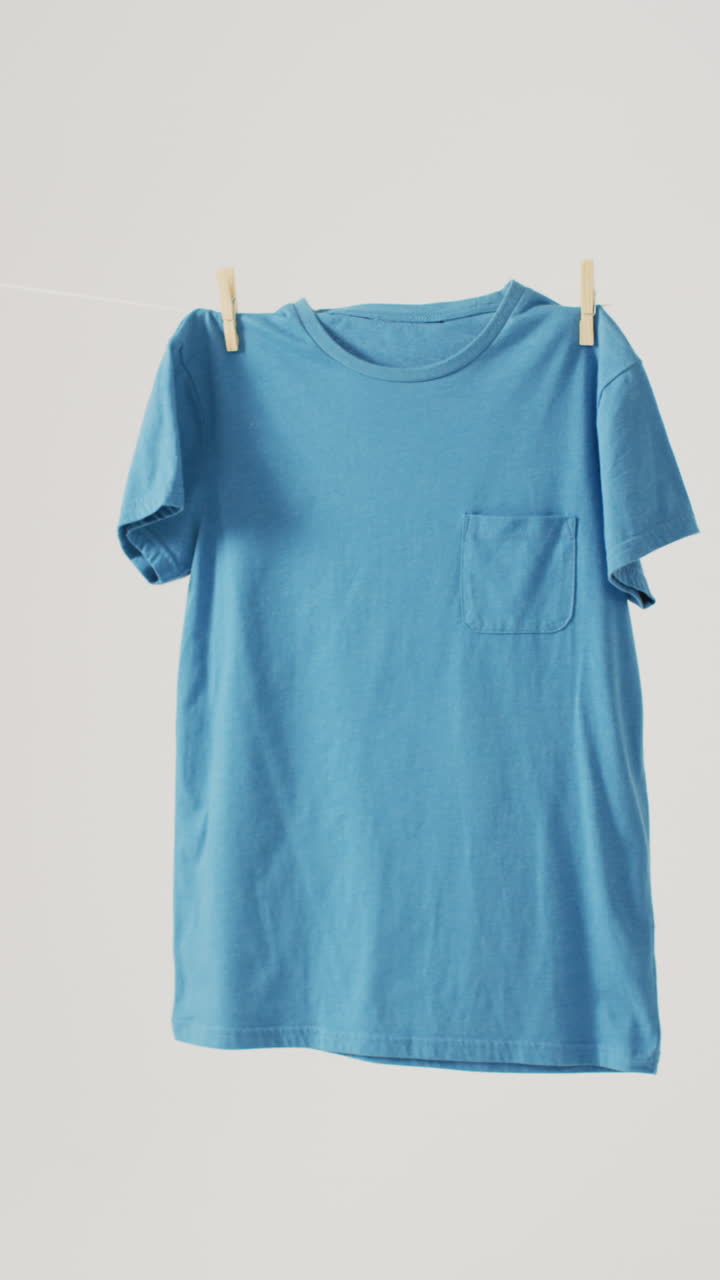 Vertical video of close up of blue t shirt hanging on white background with copy space