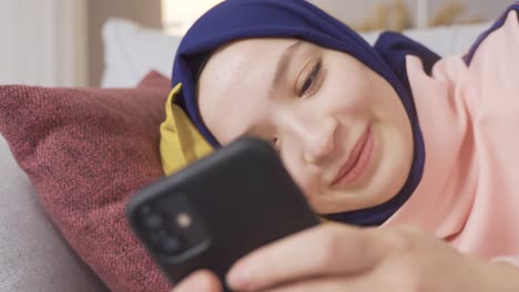 una chica musulmana satisfecha con hijab está usando un teléfono inteligente en casa.