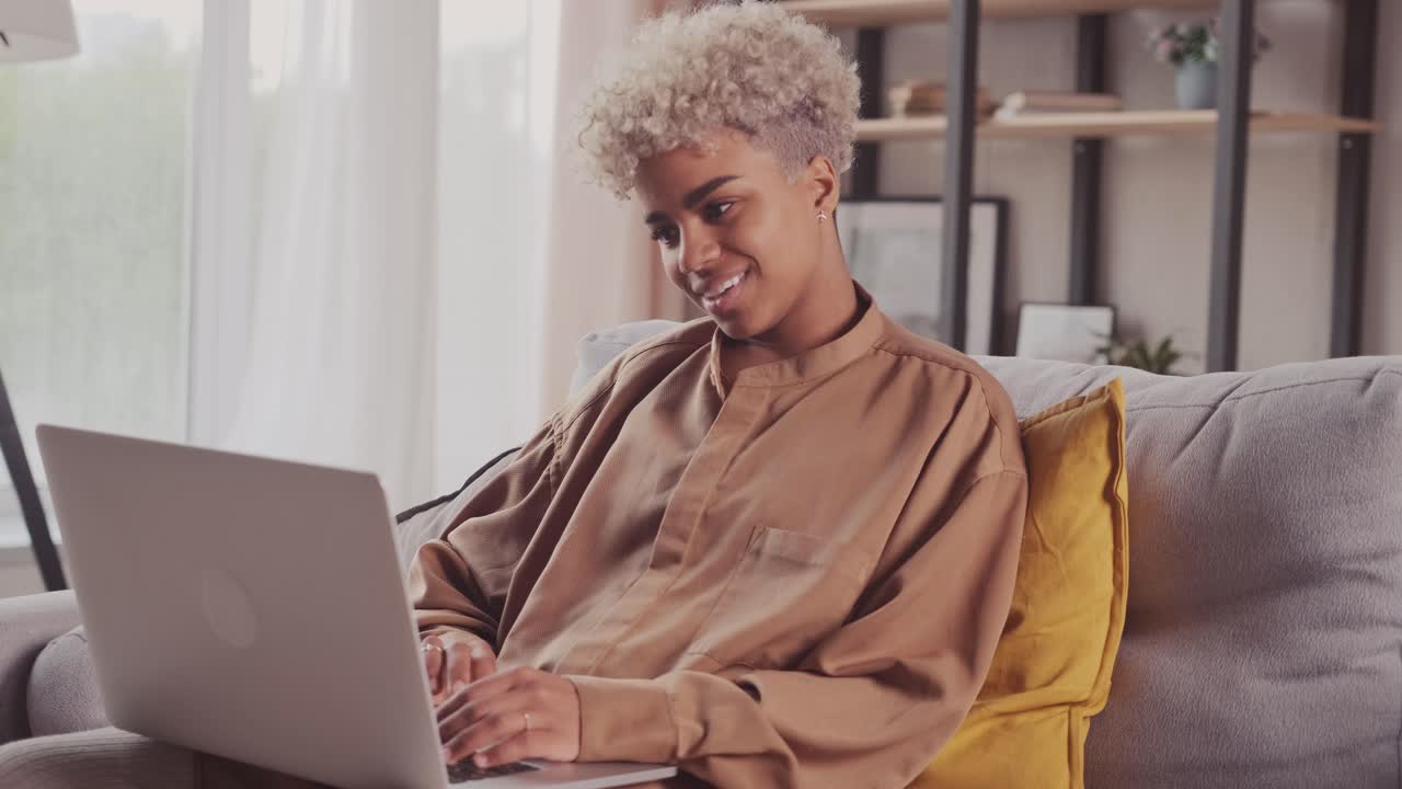 Focused african woman typing on laptop browsing internet using apps sit on sofa