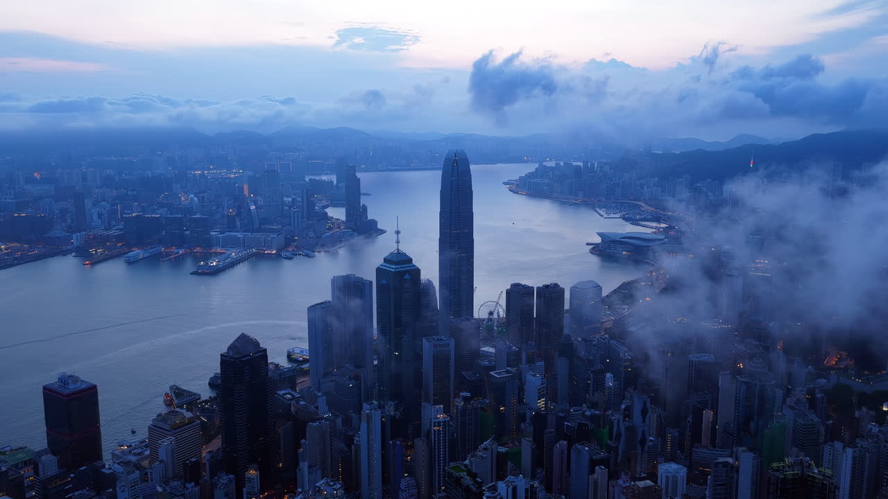 Aerial view of dense Hong Kong cityscape under foggy twilight, featuring high-rise buildings, residential towers, harbor, and mountains in the background