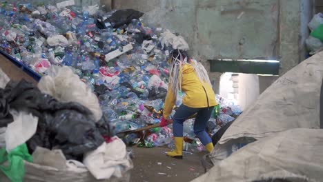 una chica con dreadlocks, en botas amarillas, cucharas botellas usadas con una pala de pato grande en una fábrica de reciclaje de plástico. enorme pila de botellas en el fondo