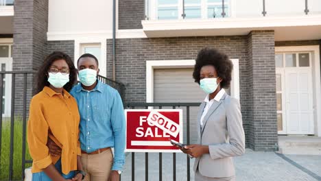 african american young happy couple in medical masks standing outdoor at new house with female real-estate agent looking at the camera