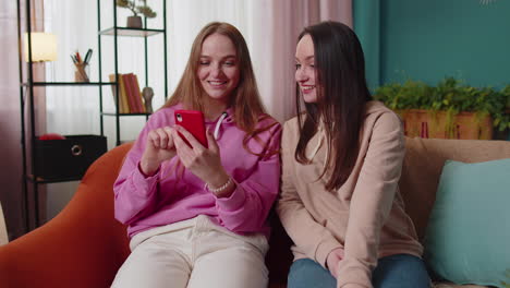 cheerful girls friends siblings watching photos on smartphone on sofa at home, enjoy social media