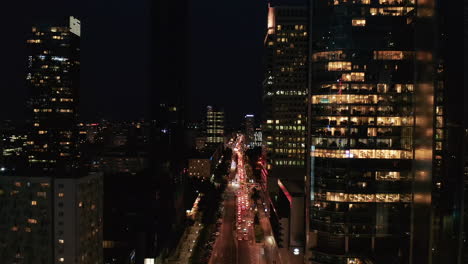 Fly-above-busy-street-at-night.-Multilane-road-lined-with-tall-office-buildings.-Traffic-jam-in-evening-rush-hour.-Warsaw,-Poland