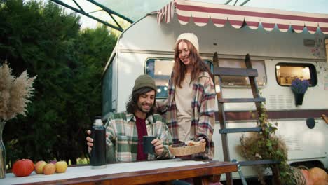 A-happy-brunette-girl-in-a-white-hat-and-a-tick-on-her-shirt-takes-out-baked-goods-for-her-boyfriend-who-pours-tea-from-a-thermos-and-sits-at-a-table-near-a-trailer-in-a-camp-outside-the-city-during-a-picnic-in-the-summer