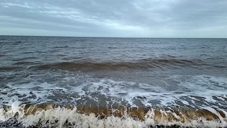 sandy dirty slow motion tide waves crashing onto stormy pebble waterfront looking out across horizon