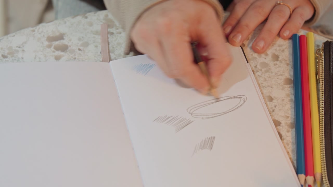 Close up of woman drawing with pencil on sketchbook creating lines and shapes. Various colored pencils arranged nearby as woman sketches focusing on hand movement and artistic process. Soft