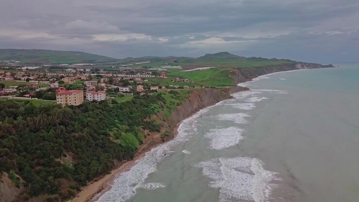 Filmato aereo della costa siciliana. Movimento in elevazione e panoramica a sinistra, rivelando la spiaggia, una zona suburbana dall'alto e un altopiano. Giornata nuvolosa con mare agitato. Colline verdi che incontrano il mare e la spiaggia sabbiosa.