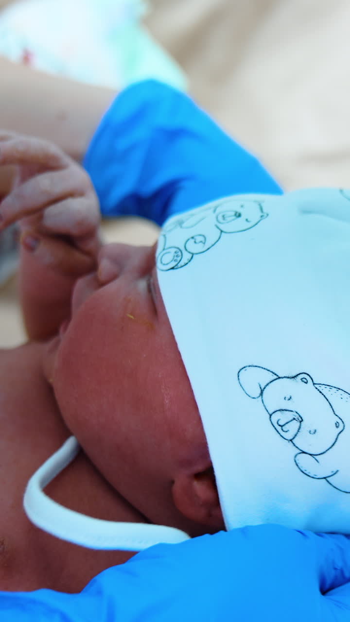 White cap with bears is put on the newborn. Gloved hands of medic tie the straps of cap while baby cries. Close up. Vertical video