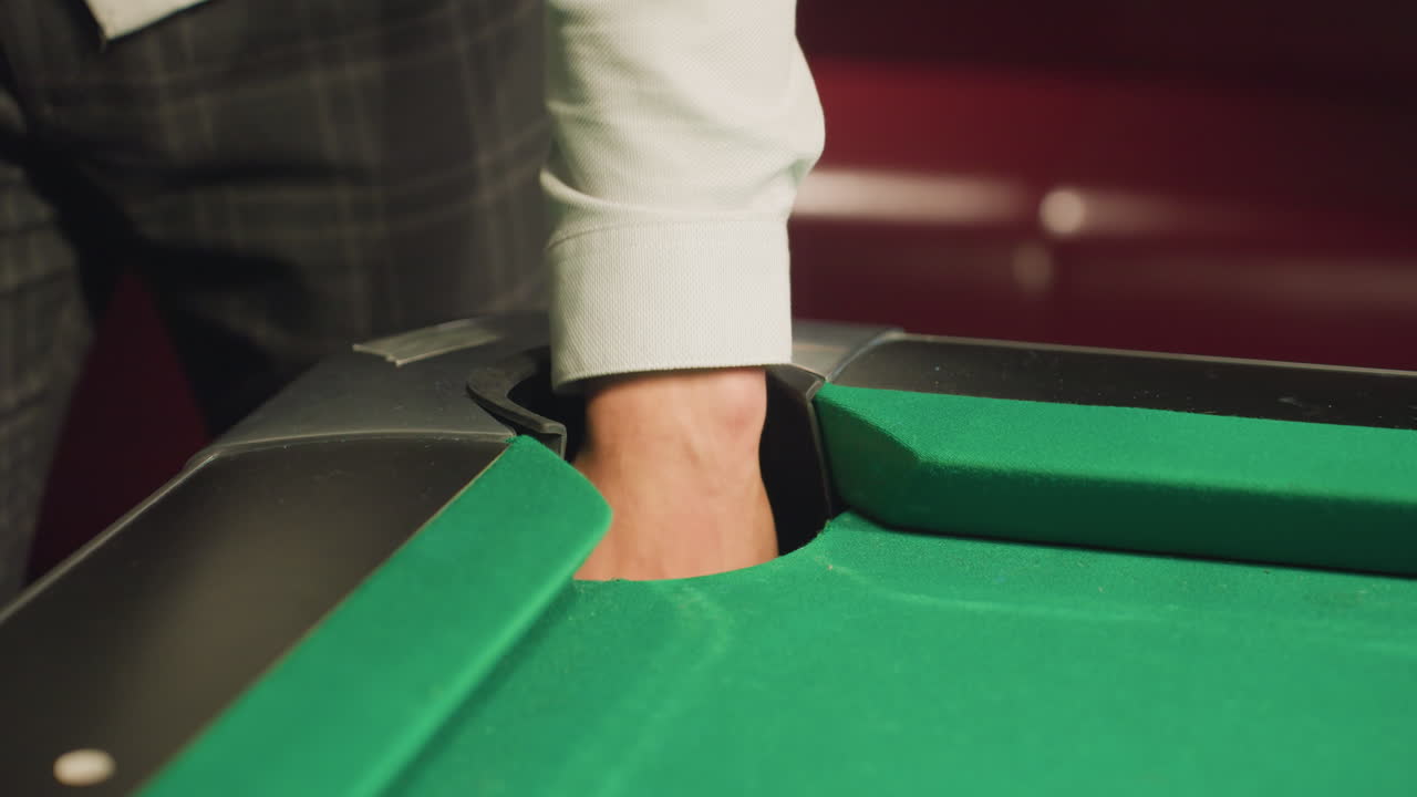 jugador de billar con camisa blanca y pantalones grises recuperando una pelota naranja del agujero de la mesa de billar verde. movimiento de la mano de primer plano e interacción del juego en un entorno escasamente iluminado con borrón de fondo suave