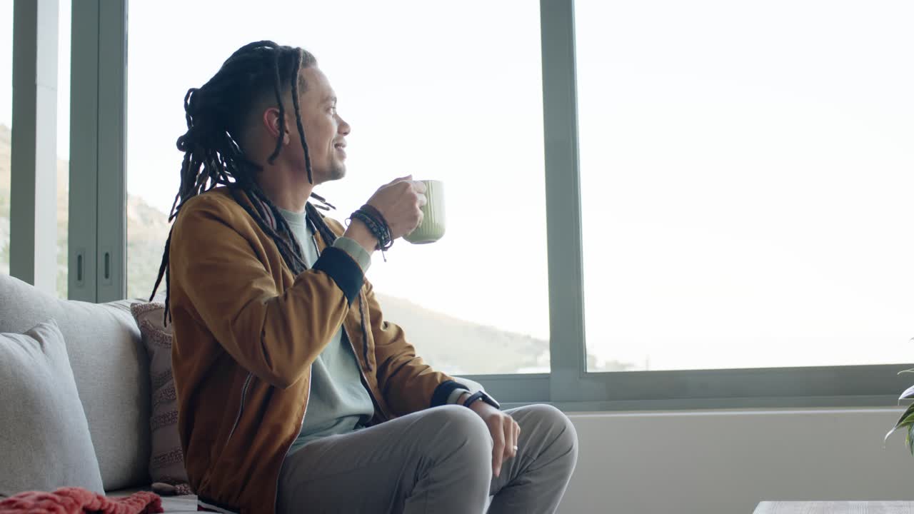 African American man relaxing on couch sipping ceramic mug and gazing out window at hillside view