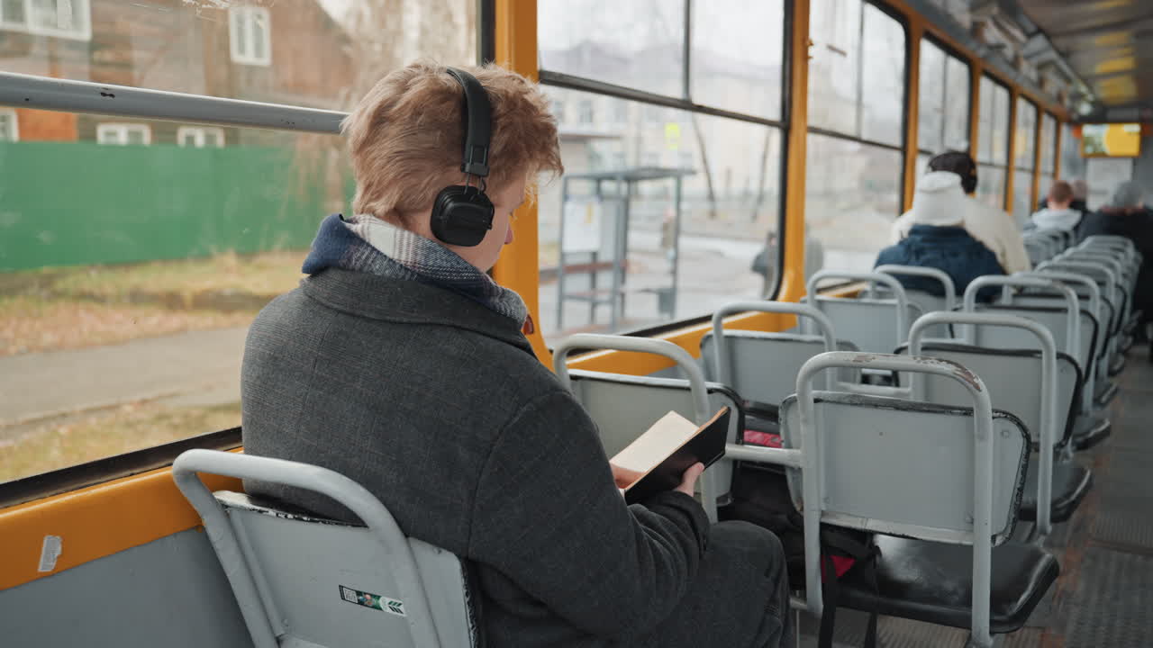 Hvid mandlig studerende og kreatør studerer i sporvogn med hovedtelefoner på, tager noter fra en bog, rygsæk ved fødderne, byblokke glider forbi uden for vinduet, flittig og fokuseret holdning, pendlerens forskningsrutine