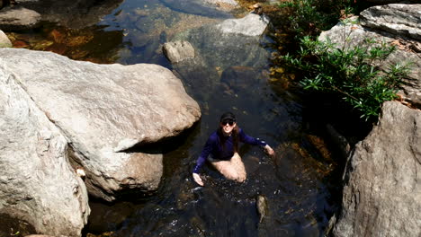 young latin woman in swimwear wading in rocky natural pool, medium down shot