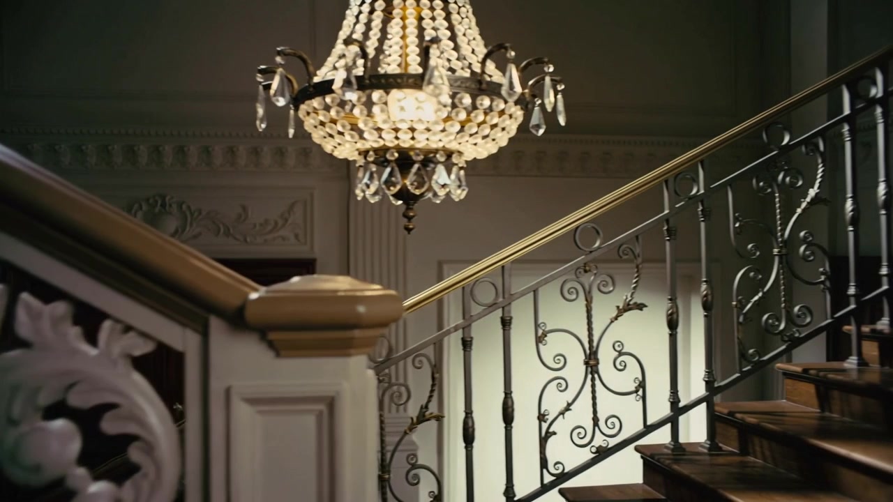 Telecamera che si muove a destra e in basso nell'atrio, rivelando una scala e un lampadario illuminato che mostra i dettagli.