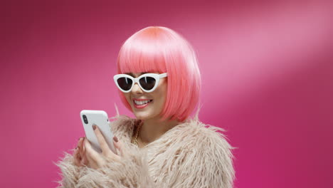 close up of cheerful young stylish woman in pink wig tapping and texting on mobile phone on colorful background and reading message 1