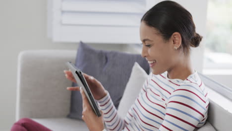 Happy-biracial-woman-sitting-on-sofa-using-tablet,-slow-motion