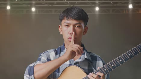 primer plano de un adolescente asiático con una guitarra mostrando un gesto de silencio mientras está de pie en el escenario
