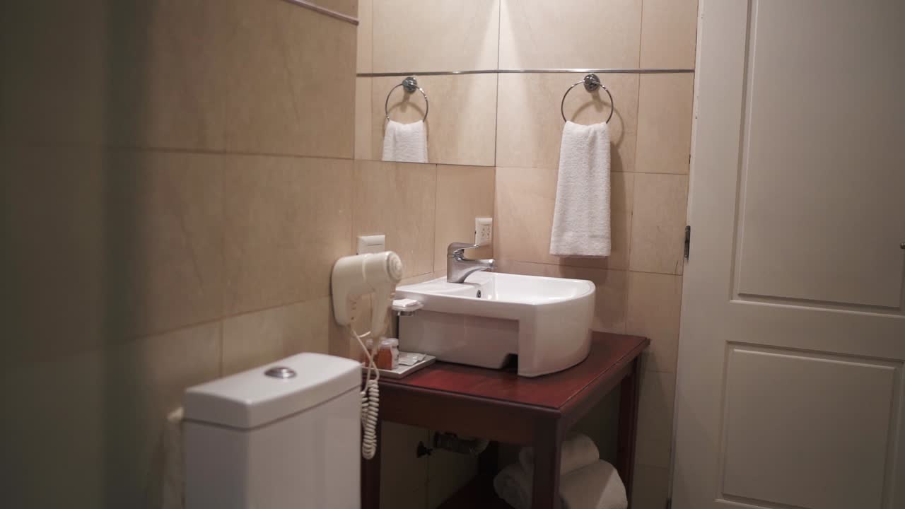 Small bathroom interior featuring a sink, toilet, hairdryer, and a white towel hanging on the wall, emphasizing hygiene and comfort in a modern hotel or apartment setting, slow motion reveal