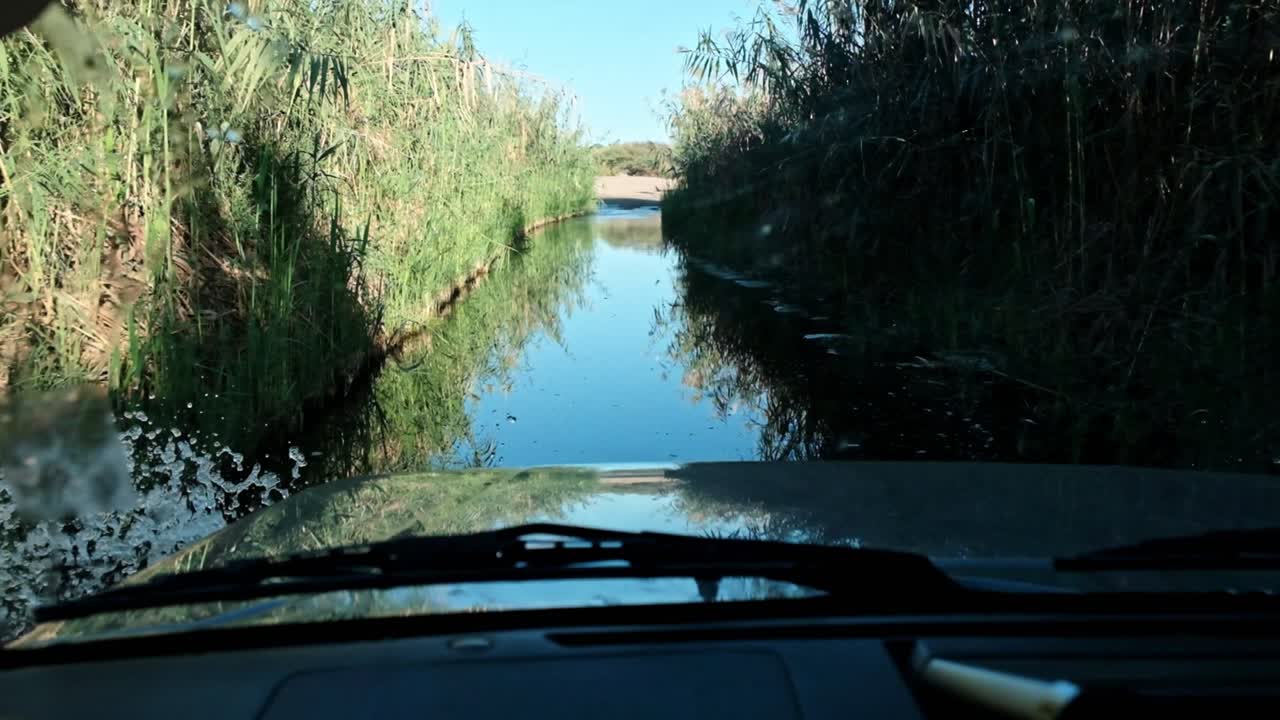 SUV vehicle driving through a flooded river crossing with reeds on either side.