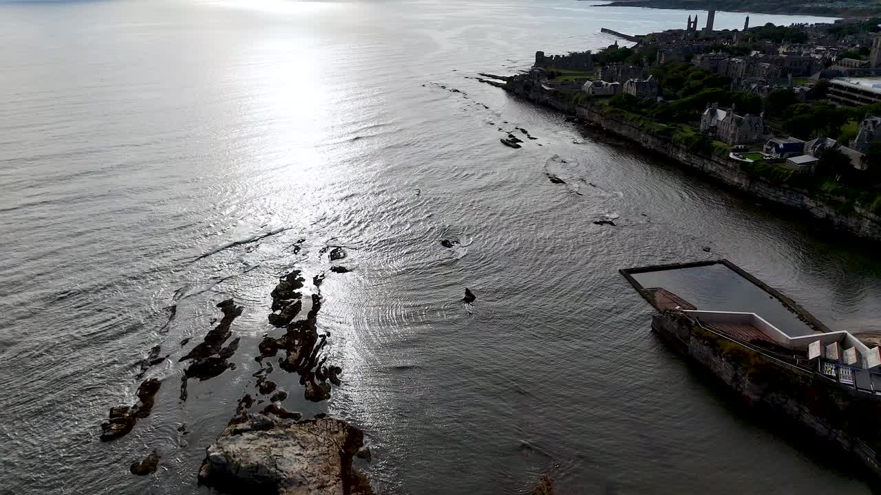 Multiple small boats move away from the rocky St Andrews shoreline under dramatic backlighting, captured in a smooth aerial tracking shot with reflective water
