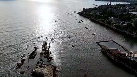 multiple small boats move away from the rocky st andrews shoreline under dramatic backlighting, captured in a smooth aerial tracking shot with reflective water