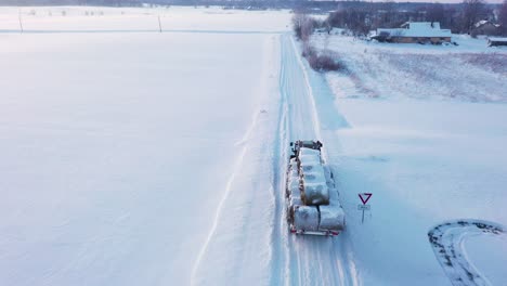 Tractor-with-haybales-driving-on-icy-road-in-winter-season,-aerial-follow-up-shot