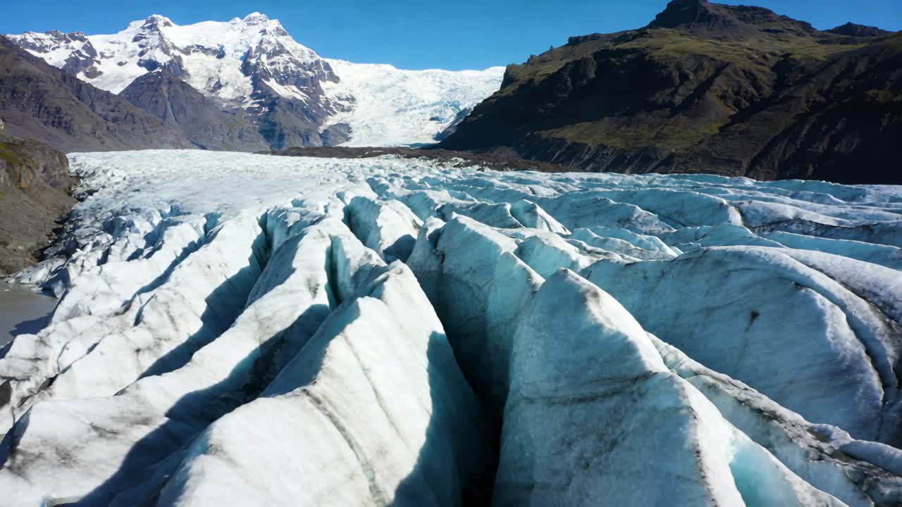 Kamera starter og beveger seg over alpinbre mot snødekte topper, viser sprekker, morene, smeltevann