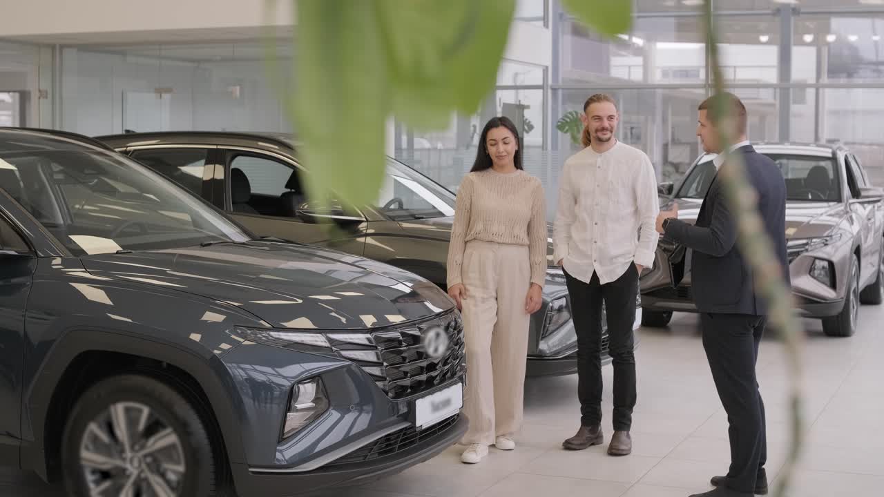 un joven vendedor de coches mostrando a una pareja de jóvenes un nuevo automóvil en el salón de concesionarios.