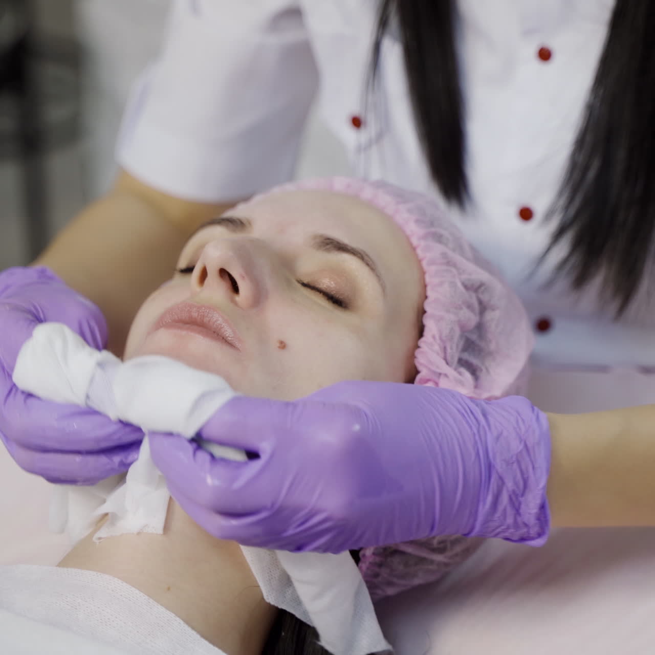 A beautician in gloves holds sanitary napkins in her both hands and cleans the skin of the client's face with it before the procedure. Close-up.