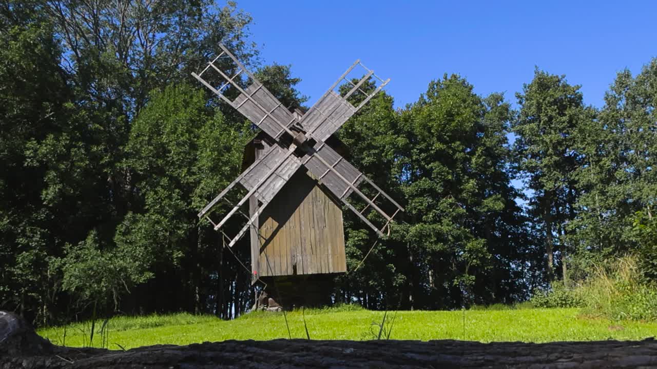 Low angle view of a traditional wooden and medieval retro windmill with large wooden paddles or blades on it at sunny summer countryside next to large trees. Blue sky in the background