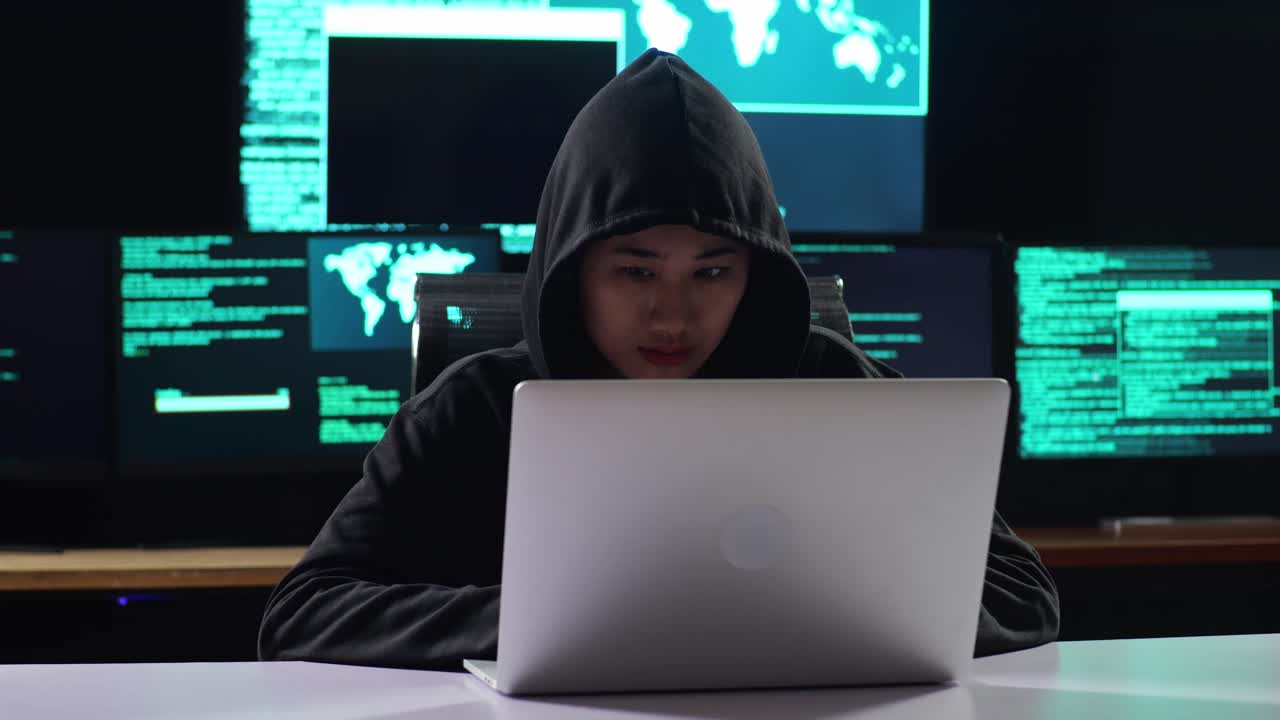 Asian Female Hacker In A Hood Works On A Laptop With Maps And Data On Display Screens Of Multiple Monitors In A Dark Office Room