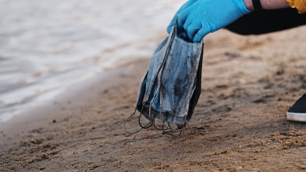 Man in medical gloves picking up a bunch of dirty medical masks from the beach of a lake. Pollution idea