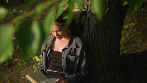 lady sits under tree reading book, leaning against trunk, blinks thoughtfully, sunlight softly illuminates her face, green leaves sway gently in breeze, partially obscuring her figure